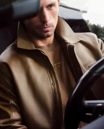 Man wearing a brown leather jacket and matching crewneck sweater seated inside a car.