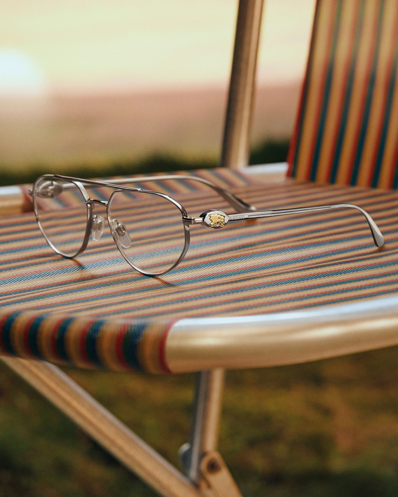 Silver aviator eyeglasses with gold logo detail resting on a striped folding chair outdoors