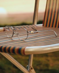 Silver aviator eyeglasses with gold logo detail resting on a striped folding chair outdoors