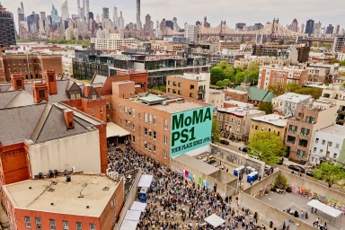 Aerial view of a large crowd gathered outside a red brick building with a teal MoMA PS1 mural in an urban setting.