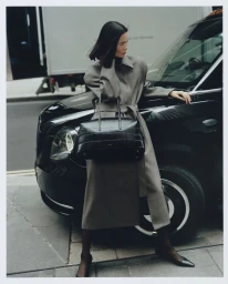 Model leaning against a black car wearing a grey belted coat and holding a black handbag
