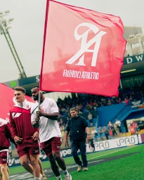 Athletes in red and white athletic wear walking across a sports field carrying large red branded flags for Ultimate Sevens.