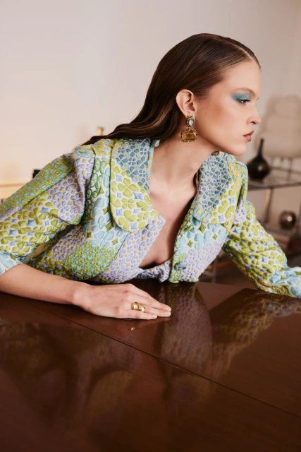 A fashionable woman wearing a patterned jacket and jewelry poses leaning on a shiny, dark wooden surface indoors.