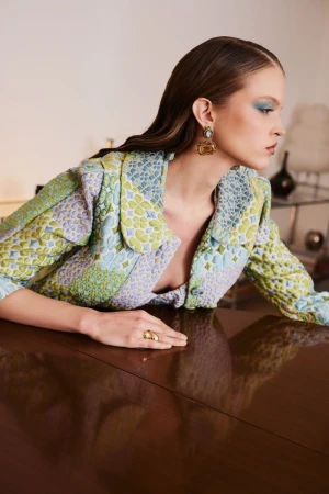 A fashionable woman wearing a patterned jacket and jewelry poses leaning on a shiny, dark wooden surface indoors.