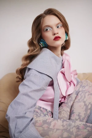 Woman with retro wavy hair wearing a pink satin top, gingham sleeve, and statement blue earrings seated on a sofa.