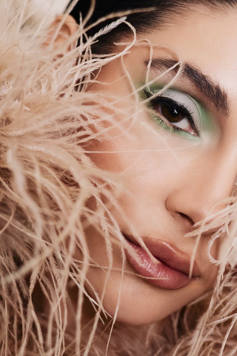 Close-up of an adult female with light skin, wearing makeup and surrounded by beige feathers in a beauty studio shot.