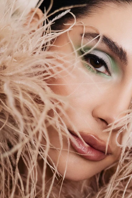 Close-up of an adult female with light skin, wearing makeup and surrounded by beige feathers in a beauty studio shot.