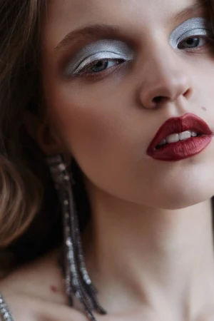 Close-up beauty portrait of a woman featuring bold metallic silver eyeshadow, dark red lipstick, and long crystal drop earrings.