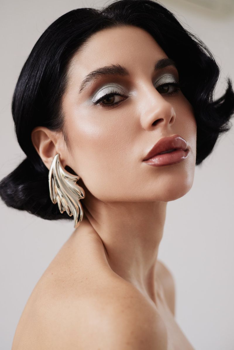 Elegant closeup portrait of a young adult woman with dark hair, silver eye shadow, a large earring, and bare shoulders, indoors.