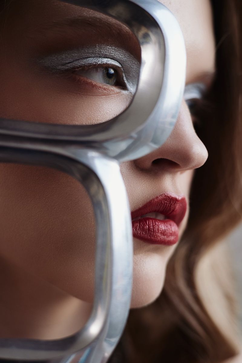 Close-up portrait of a woman with silver eye makeup and red lipstick, partially obscured by a silver metal frame, showcasing beauty and fashion.