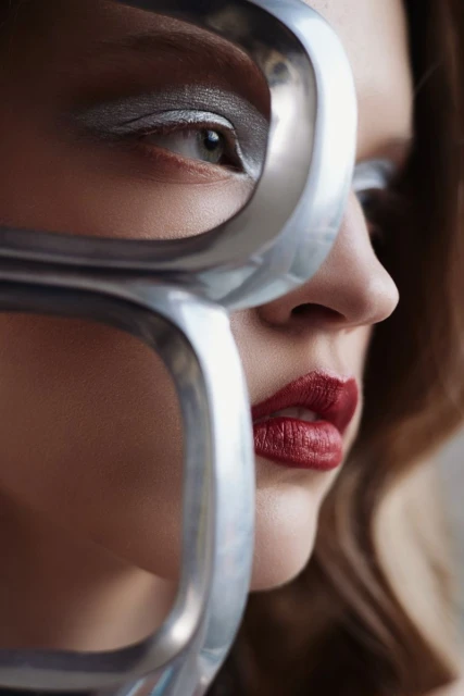 Close-up portrait of a woman with silver eye makeup and red lipstick, partially obscured by a silver metal frame, showcasing beauty and fashion.
