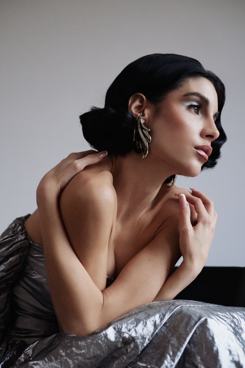 Woman with retro black wavy hair wearing a sculptural gold earring and a textured metallic silver dress.