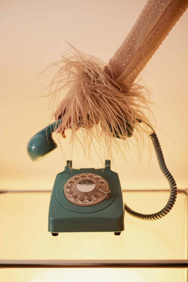 Vintage rotary phone with receiver resting on the fluffy feather-trimmed arm of a woman's dress, set against a soft, diffused light.