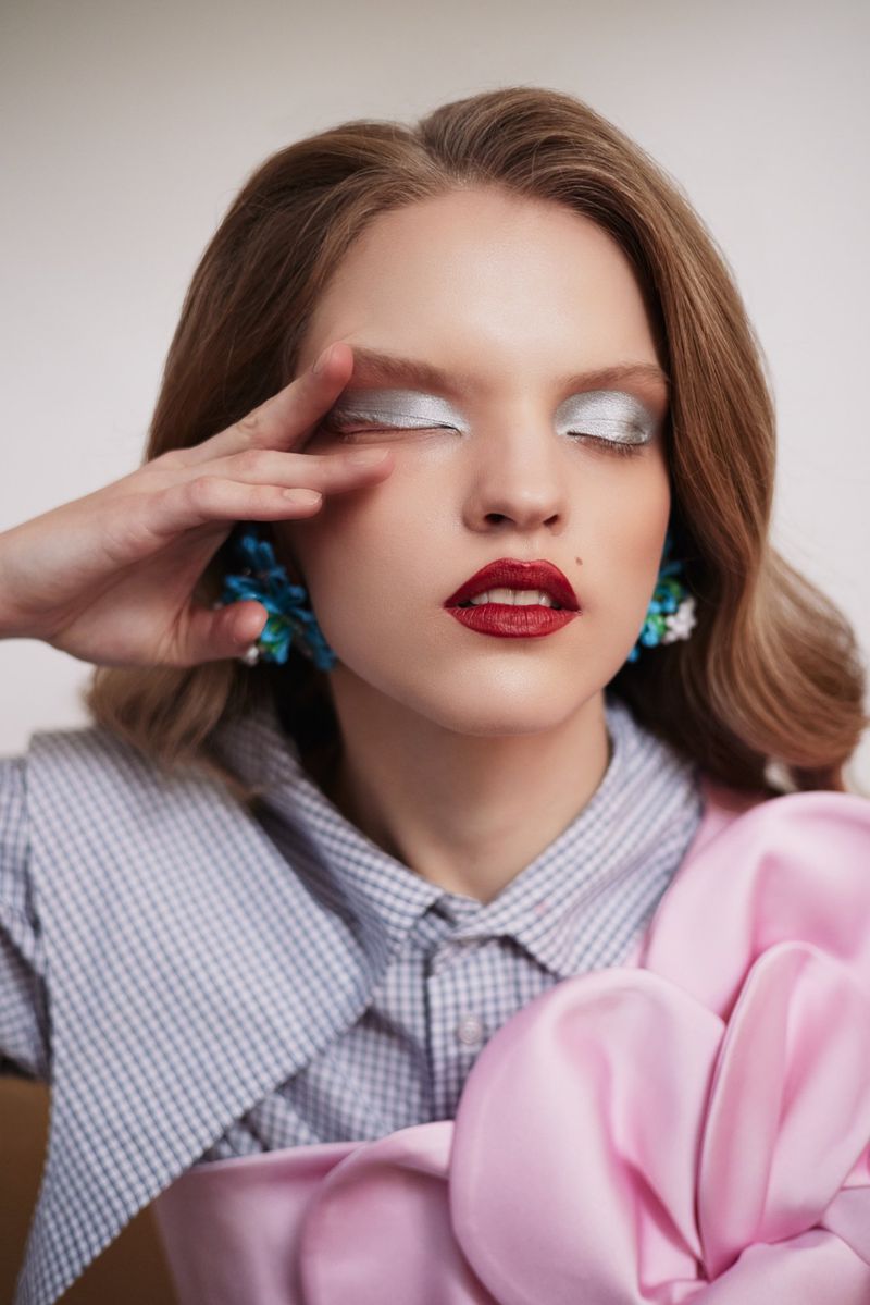 Elegant woman with silver eyeshadow and red lipstick, posing in a light blue and white checkered blouse and a pink fabric flower.