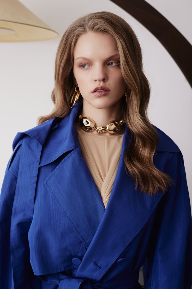 Adult woman in a studio setting wears a vibrant blue trench coat and a gold choker necklace, showcasing contemporary fashion.
