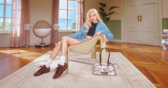 ROSÉ seated in a modern armchair wearing a blue jacket and brown sneakers with a Puma handbag on the floor