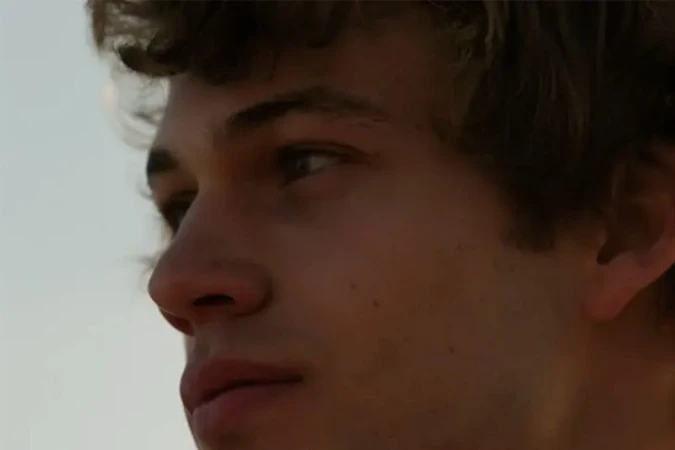 Close-up portrait of a young adult male with light skin and light brown curly hair, looking off-camera with a serious expression.