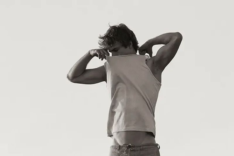 Monochrome portrait of a shirtless adult male in jeans, pulling a tank top over his head, with a light, neutral backdrop.