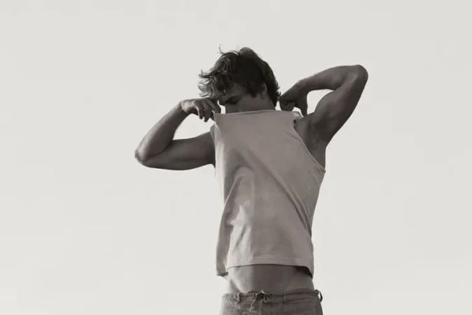 Monochrome portrait of a shirtless adult male in jeans, pulling a tank top over his head, with a light, neutral backdrop.