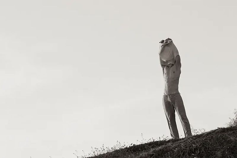 Black and white outdoor shot of adult male removing his t-shirt on a grassy hill against a bright sky backdrop, shirtless and wearing pants.