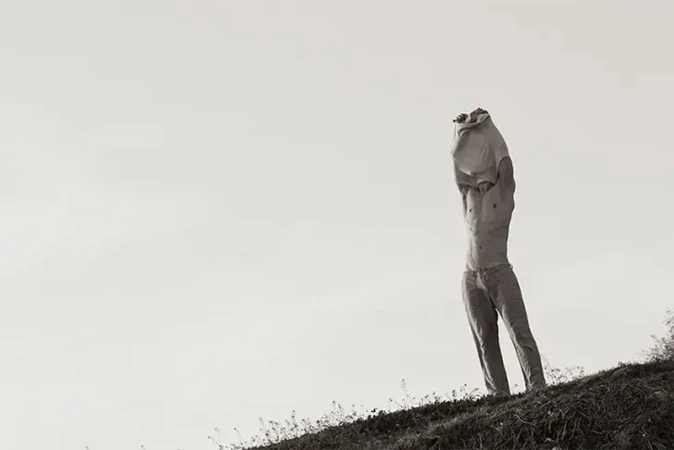 Black and white outdoor shot of adult male removing his t-shirt on a grassy hill against a bright sky backdrop, shirtless and wearing pants.