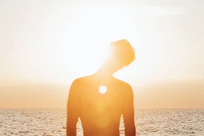 Backlit silhouette of an adult with a blurred background of the ocean under a bright golden sky with sun flares during daytime.