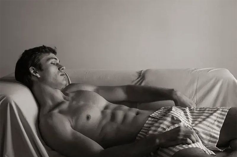 Shirtless young man with eyes closed resting back on a draped sofa wearing striped boxer shorts.