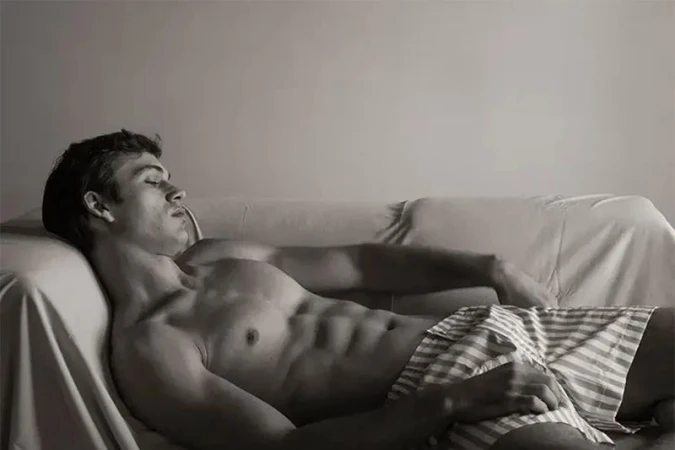 Shirtless young man with eyes closed resting back on a draped sofa wearing striped boxer shorts.