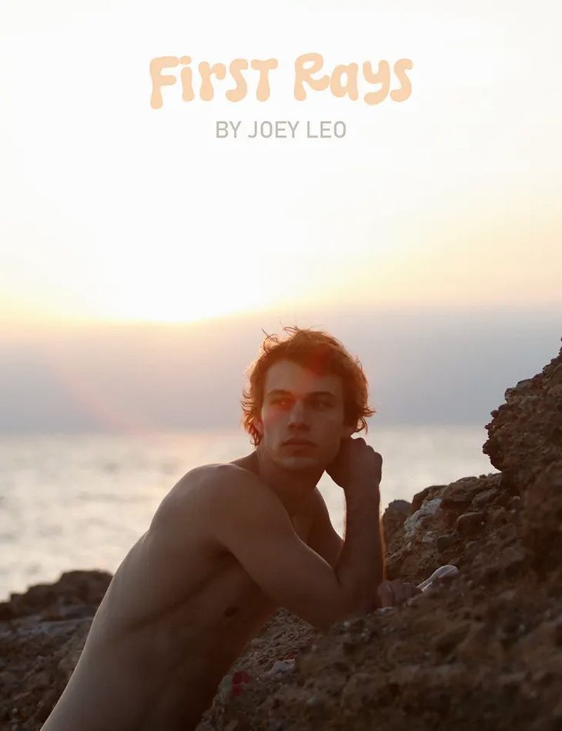 Young adult shirtless male poses on rocks near the ocean at sunrise with orange light, creating a portrait with sea background.