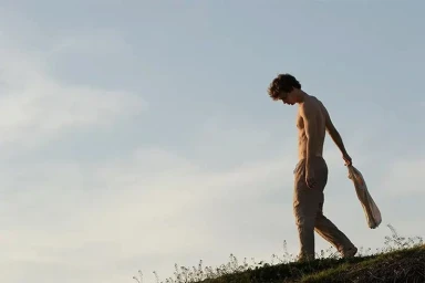 Shirtless young man walking outdoors against a pale sky holding a garment