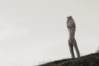 A man pulls a light-colored shirt over his head while standing on a grassy hillside against a pale sky.