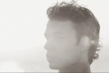 Close-up profile portrait of a young man with tousled hair against a bright, overexposed light background.