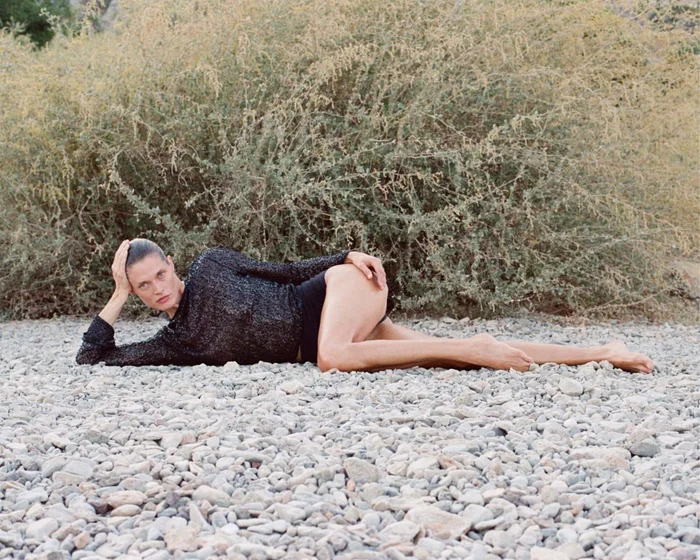 Model lying on a rocky ground wearing a dark textured top against sparse foliage
