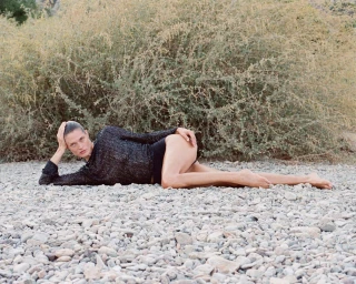 Model lying on a rocky ground wearing a dark textured top against sparse foliage