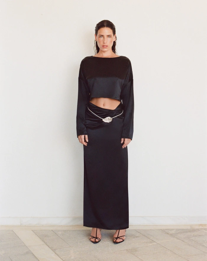 Model standing against a plain wall wearing a black long-sleeve crop top and matching long skirt with a decorative belt.