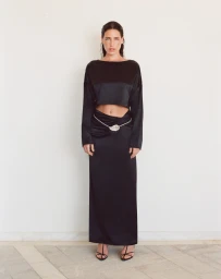 Model standing against a plain wall wearing a black long-sleeve crop top and matching long skirt with a decorative belt.
