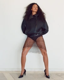 Model with dark curly hair wearing an oversized black leather jacket and fishnet overlay against a pale wall.