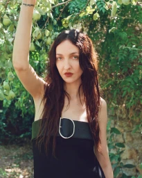 Model with long dark hair raising her arm among green foliage for SIR. Resort 2027 campaign.