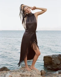 Model standing on coastal rocks in Greece wearing a dark fringed satin dress with arms raised against the sea.