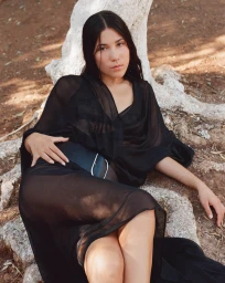 Model in a black sheer dress leaning against a whitewashed tree trunk on earthy ground