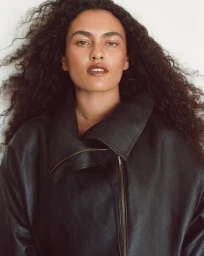 Portrait of a woman with long curly dark hair wearing a black leather jacket against a light background.