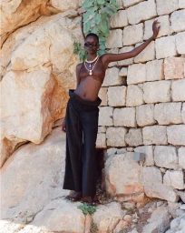 Model standing outdoors leaning against a weathered stone wall wearing dark trousers and layered necklaces