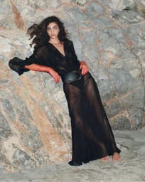 Model in sheer black dress and red gloves leaning against a rocky stone wall in Greece