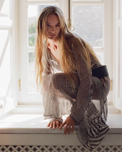 Model in a striped dress crouching on a windowsill against a bright window for the SIR. Resort 2027 campaign.