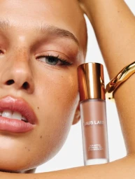 Close-up portrait of model holding a Haus Labs bronzing product bottle near her face against a white background