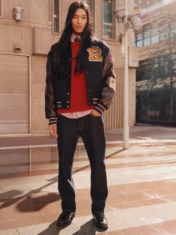 Model with long dark hair wearing a varsity jacket, red sweater, and dark jeans outdoors on a paved street.