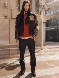 Model with long dark hair wearing a varsity jacket, red sweater, and dark jeans outdoors on a paved street.