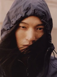 Close-up portrait of a model with long dark hair wearing a hooded navy blue jacket for H&M Fall 2026 campaign.