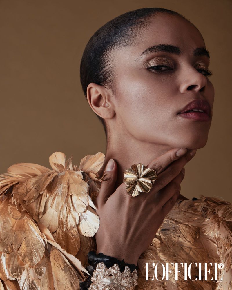 Fashion editorial headshot of a Black woman wearing a feathered dress and bold gold jewellery, posing against a neutral backdrop indoors.