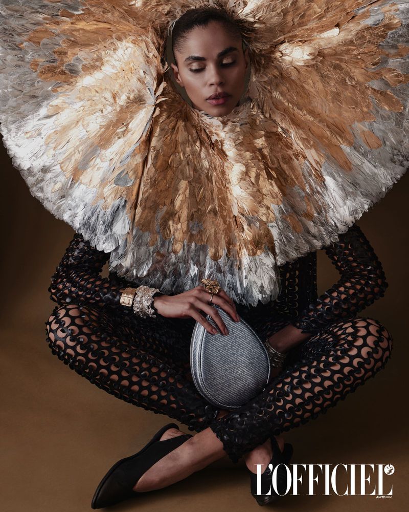A woman in elaborate feather collar and black patterned bodysuit poses cross-legged with a silver clutch in a studio setting with gold background.